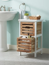 A white two-drawer chest with wooden slatted drawers, placed next to a sink in a light blue bathroom. The top of the chest features a small basket and a decorative item, while one drawer is partially open, revealing its interior. The floor is light-colored, complementing the overall aesthetic