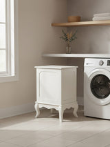 Ivory finish laundry hamper with a lift-up lid, featuring decorative legs and a smooth surface, positioned next to a washing machine in a bright, modern laundry room with a light-colored wall and wooden shelf above