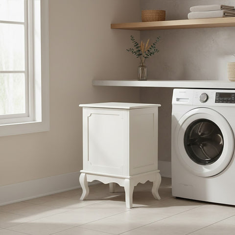 Ivory finish laundry hamper with a lift-up lid, featuring decorative paneling and elegant curved legs, positioned next to a washing machine in a bright laundry room with natural light streaming through a window and a wooden shelf above