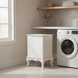 Ivory finish laundry hamper with a lift-up lid, featuring decorative paneling and elegant curved legs, positioned next to a washing machine in a bright laundry room with natural light streaming through a window and a wooden shelf above