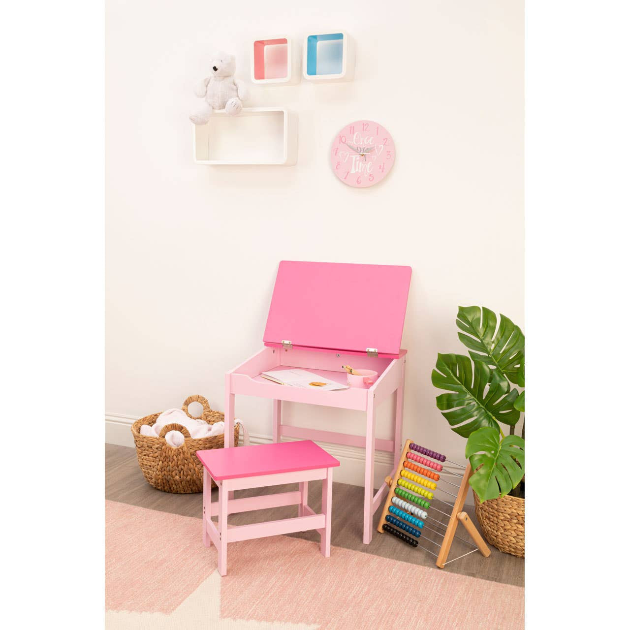 Three white floating wall cubes arranged in a staggered formation on a light-colored wall. The cubes are empty, showcasing their open design. A pink chair and a small pink stool are positioned nearby on a soft rug, with a colorful stack of books and a potted plant visible in the corner. Decorative wall art and a clock are also displayed above the cubes