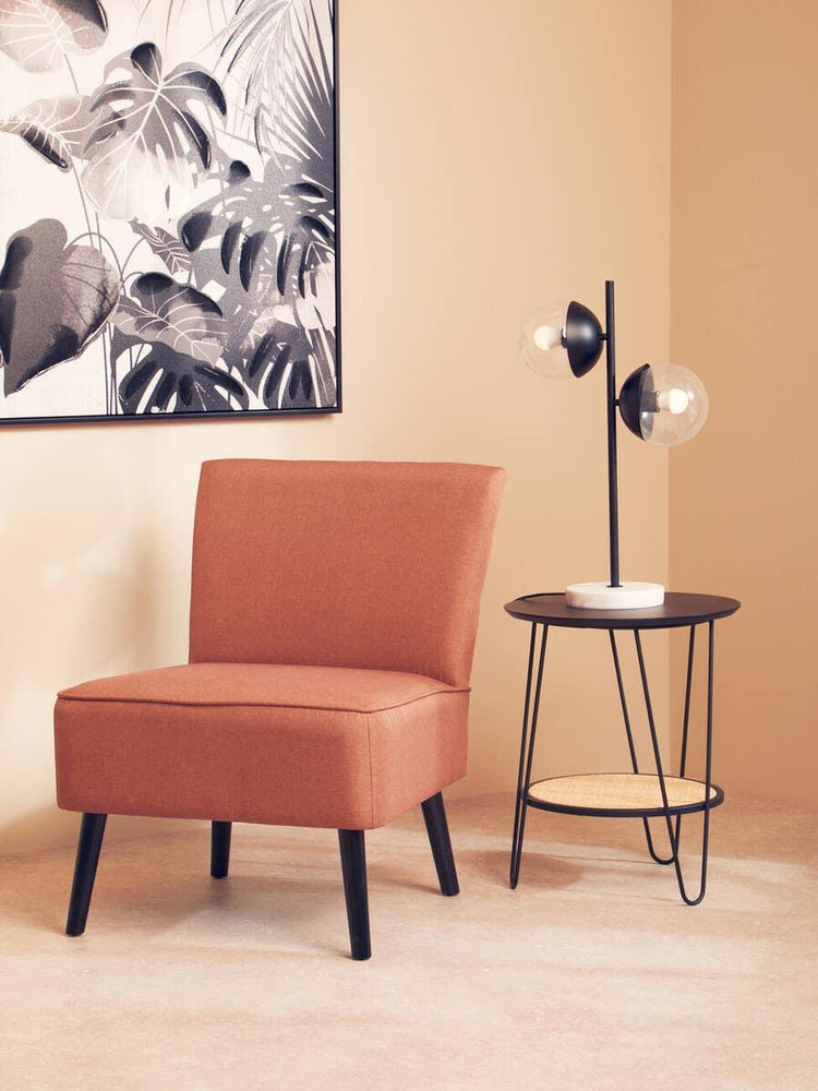 Rust accent chair with black legs beside round side table and modern table lamp