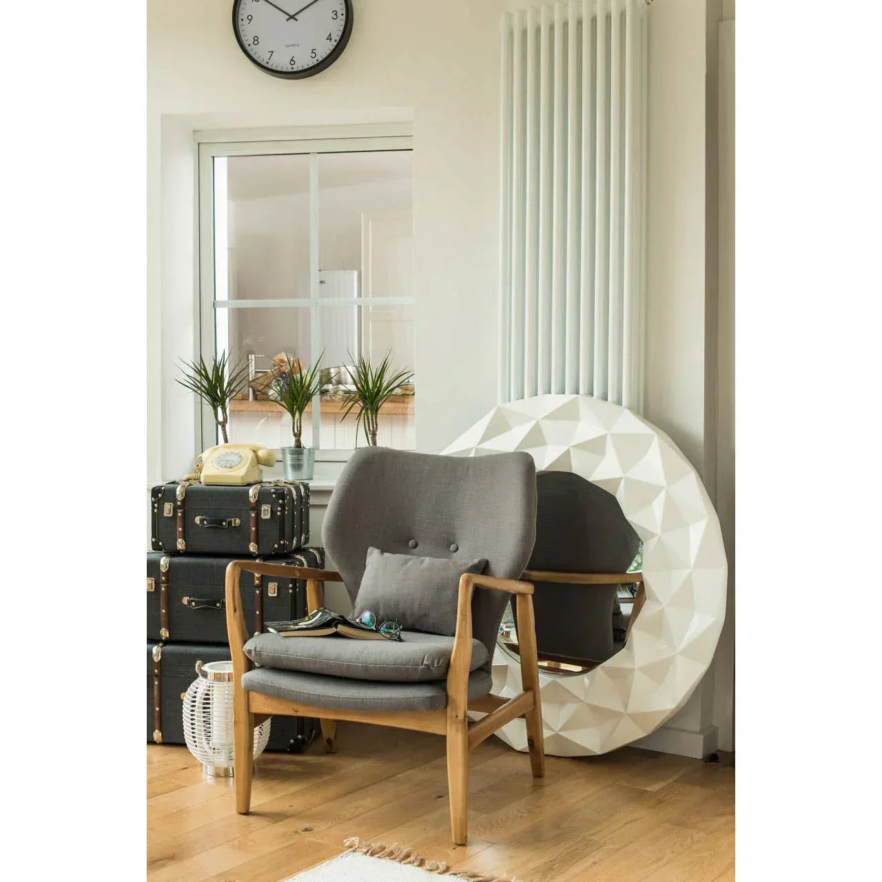 Grey accent chair with wooden frame beside stacked suitcases, round mirror and potted plants
