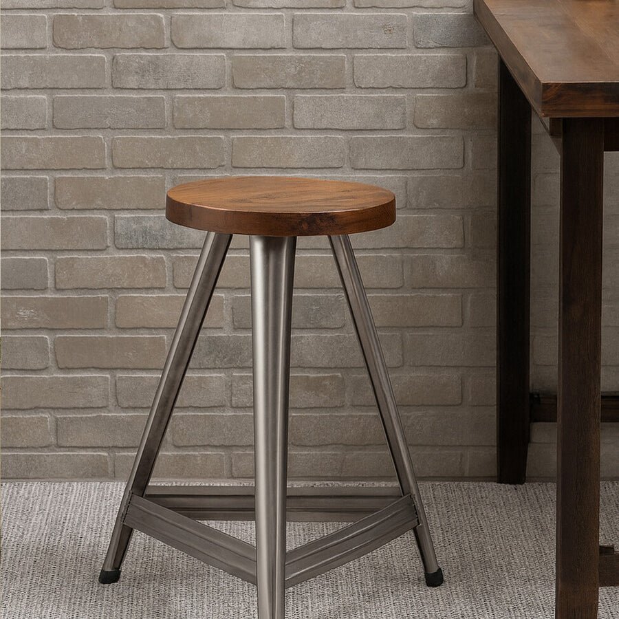 Large wooden stool featuring a round top with a warm wood finish, supported by a sturdy base in an antique silver tone. The stool has three legs that form a stable triangular shape, set against a textured light-colored wall and a wooden table in the background