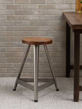 Large wooden stool featuring a round top with a warm wood finish, supported by a sturdy base in an antique silver tone. The stool has three legs that form a stable triangular shape, set against a textured light-colored wall and a wooden table in the background