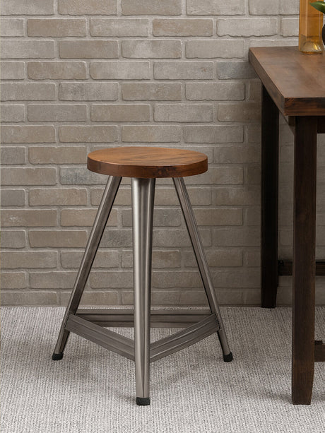 Large wooden stool featuring a round top with a warm wood finish, supported by a sturdy base in an antique silver tone. The stool has three legs that form a stable triangular shape, set against a textured light-colored wall and a wooden table in the background