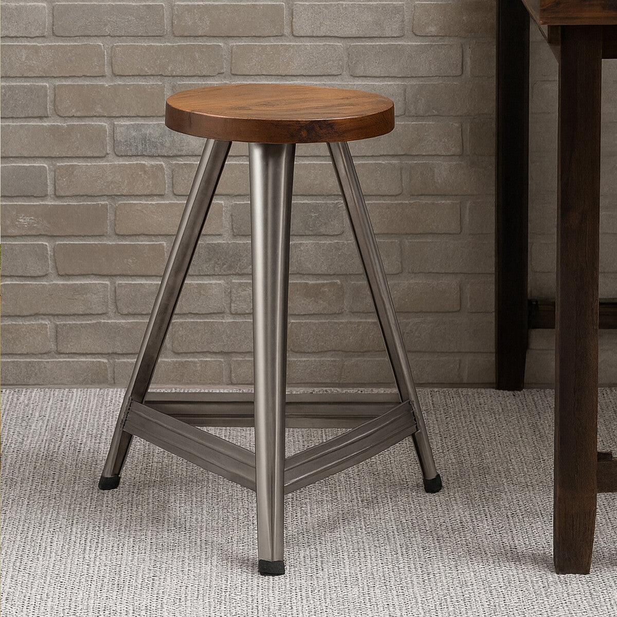 Large wooden stool featuring a round top with a warm wood finish and a sturdy base in an antique silver finish. The base has a triangular design with three legs, providing stability. The stool is placed on a light-colored carpet against a textured wall