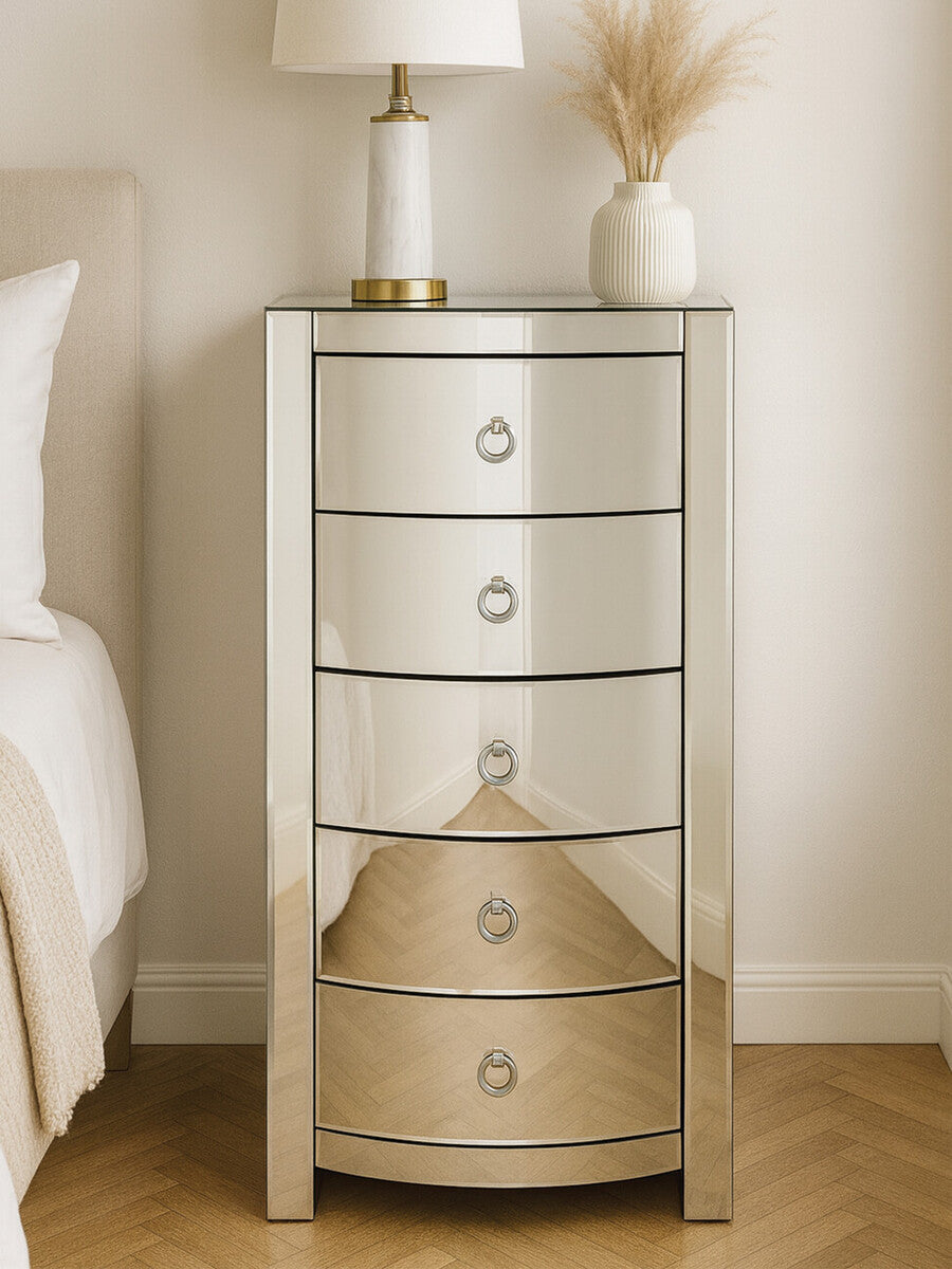 Curved tall boy drawers with a mirrored finish, featuring four spacious drawers with round handles, placed beside a bed with a light-colored duvet and a lamp on top, against a neutral wall and wooden floor