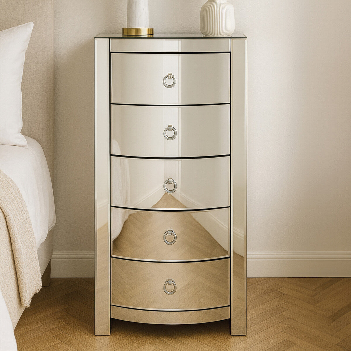 Verona Curved Tall Boy Drawers featuring a sleek mirrored finish, five curved drawers with circular handles, and a stylish design that enhances modern home decor. The unit stands on a wooden floor beside a light-colored bed with neutral bedding