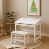A white children's desk with a slanted top and a matching stool, placed on a circular rug in a cozy room. A framed artwork featuring a green tree is hung on the wall, and a small plant sits on a side table next to the desk. A stuffed animal is visible on the floor