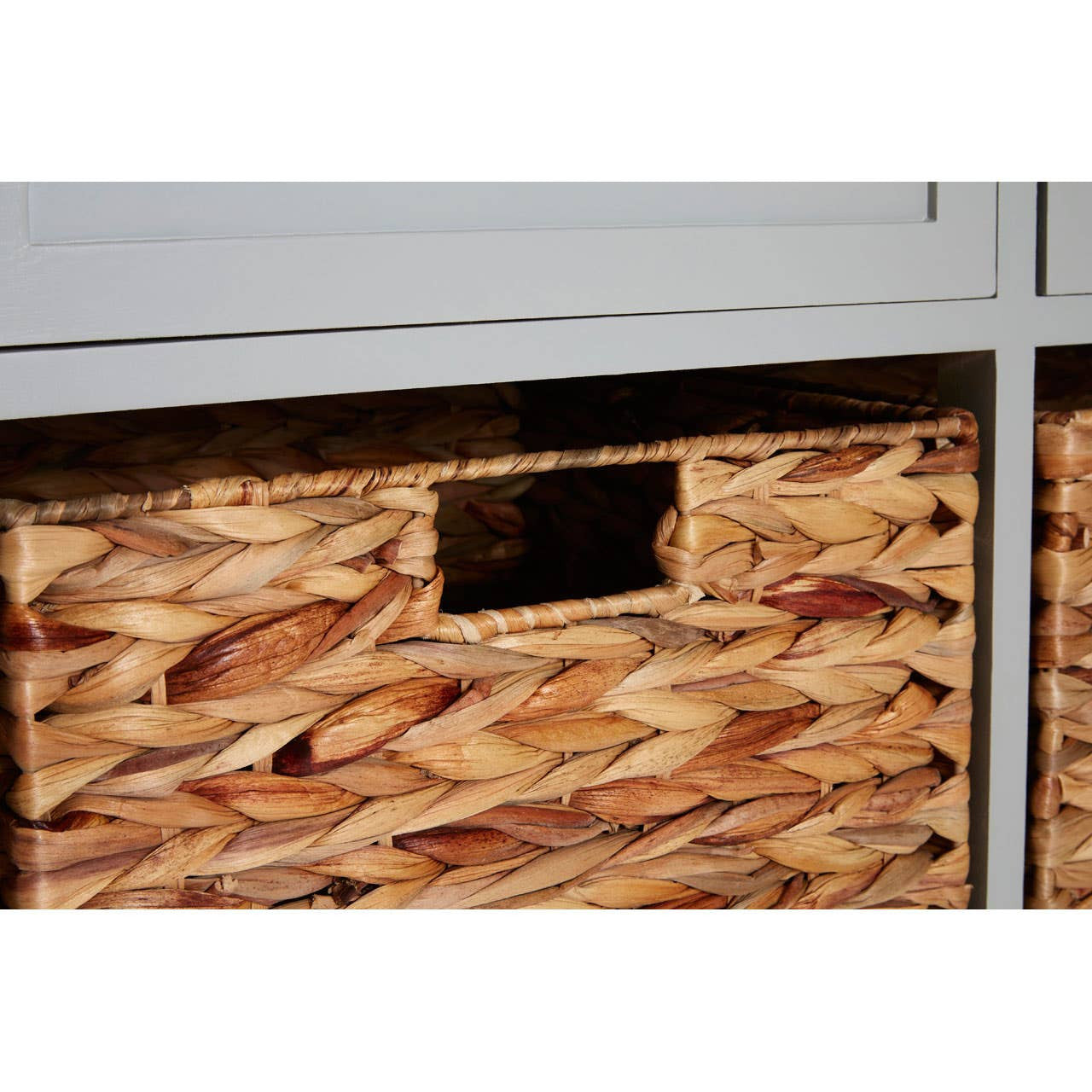 Close-up view of a woven basket with a cut-out handle, part of the Vermont Two Drawers Four Baskets Cabinet, showcasing the natural texture and design of the basket. The cabinet features a light gray finish with visible drawer fronts above the baskets