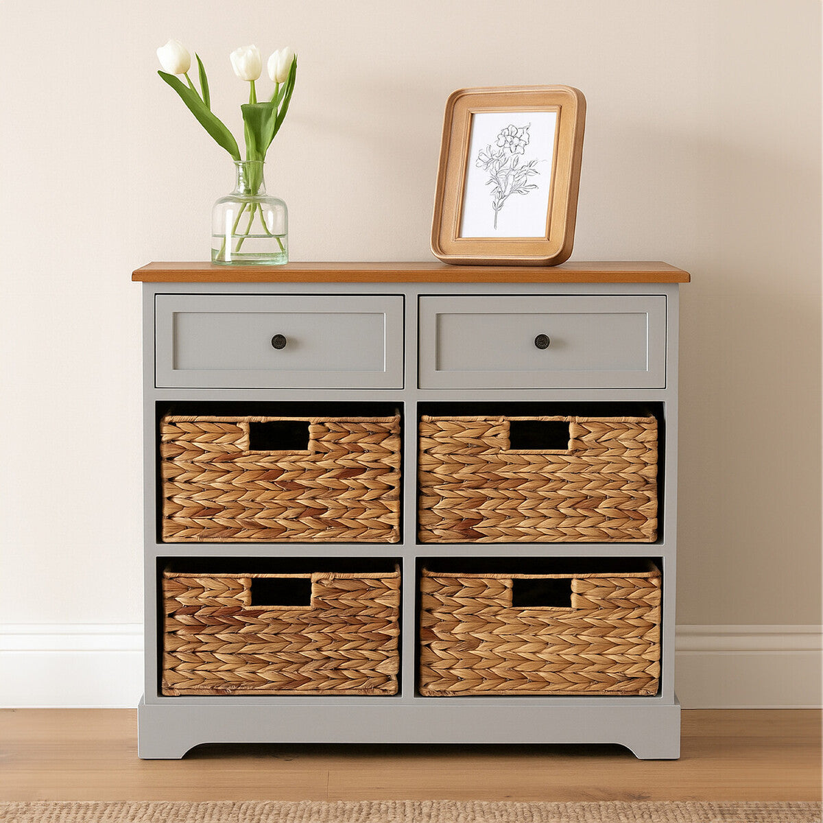 A light gray cabinet with a wooden top featuring two drawers and four woven baskets. The cabinet is positioned against a neutral wall, with a small vase of white flowers and a framed picture placed on top. The floor is wooden, adding warmth to the setting
