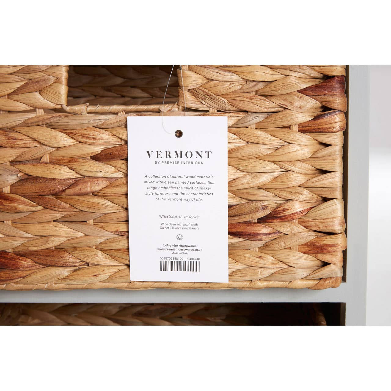 The Vermont Two Drawers Four Baskets Cabinet features a natural woven texture on the baskets, with a label attached that includes product information. The cabinet has a sleek design with a light-colored frame, showcasing the contrast between the woven baskets and the cabinet structure