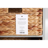The Vermont Two Drawers Four Baskets Cabinet features a natural woven texture on the baskets, with a label attached that includes product information. The cabinet has a sleek design with a light-colored frame, showcasing the contrast between the woven baskets and the cabinet structure
