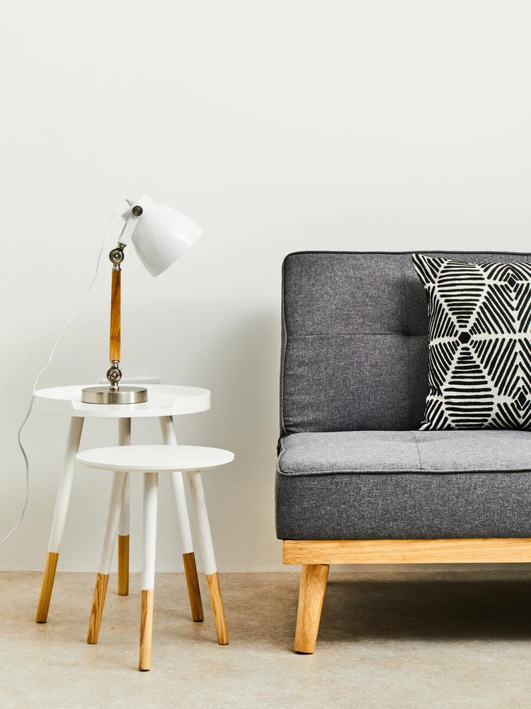 Round side table with a white top and natural wood legs, positioned next to a gray sofa with a patterned cushion. A modern lamp with a white shade is placed on the table, and the background features a plain white wall. The floor is covered with a light-colored rug