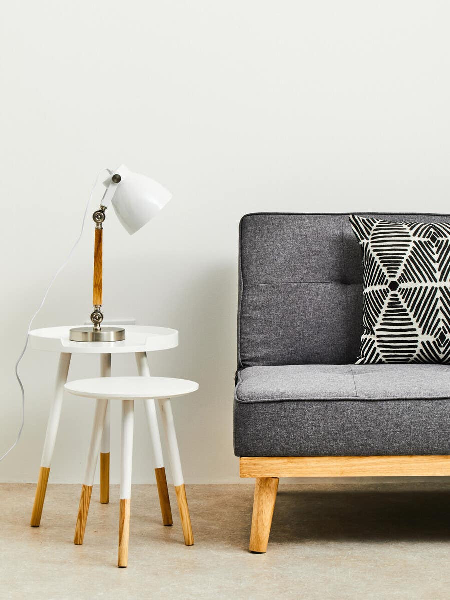 Round side table with a white top and natural wood legs, positioned next to a gray sofa with a patterned cushion. A modern lamp with a white shade is placed on the table, and the background features a plain white wall. The floor is covered with a light-colored rug