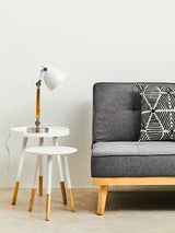 Round side table with a white top and natural wood legs, positioned next to a gray sofa with a patterned cushion. A modern lamp with a white shade is placed on the table, and the background features a plain white wall. The floor is covered with a light-colored rug