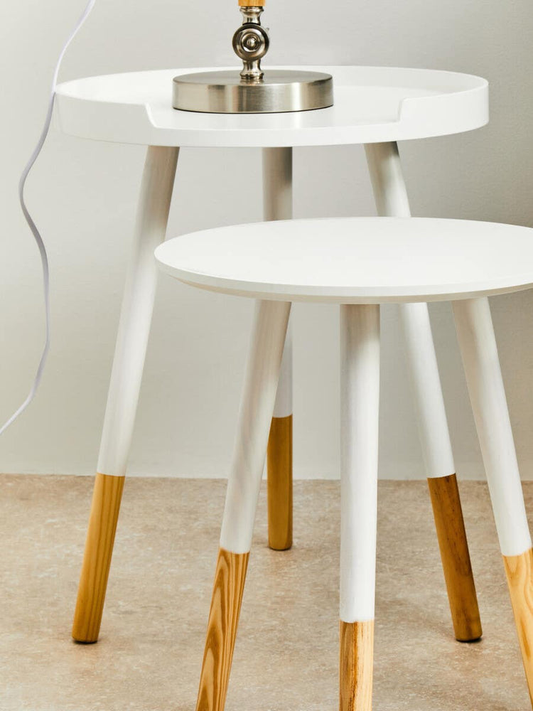 Round side table featuring a white top and natural wood legs, with two tiers. The legs are tapered and have a natural wood finish, while the tabletop is smooth and circular. A small decorative item is placed on the top surface, and the table is positioned on a light-colored floor