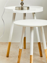 Round side table featuring a white top and natural wood legs, with two tiers. The legs are tapered and have a natural wood finish, while the tabletop is smooth and circular. A small decorative item is placed on the top surface, and the table is positioned on a light-colored floor