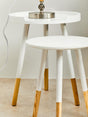 Round side table featuring a white top and natural wood legs, with two tiers. The legs are tapered and have a natural wood finish, while the tabletop is smooth and circular. A small decorative item is placed on the top surface, and the table is positioned on a light-colored floor