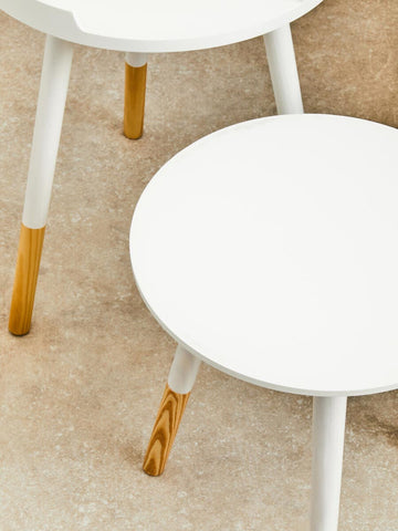 Round side table with a white top and natural wood legs, featuring a minimalist design. The legs are tapered and have a light wood finish, contrasting with the smooth white surface. The table is positioned on a neutral-colored floor, enhancing its modern aesthetic