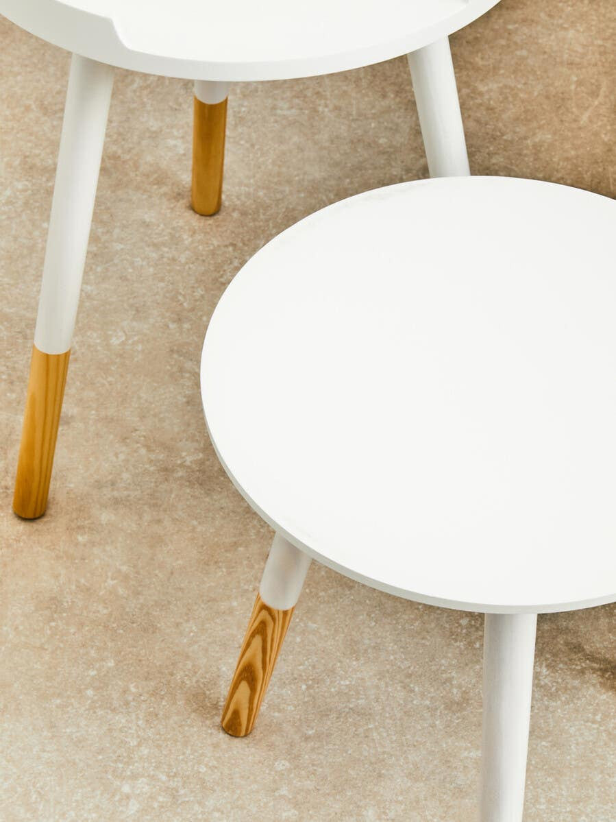 Round side table with a white top and natural wood legs, featuring a minimalist design. The legs are tapered and have a light wood finish, contrasting with the smooth white surface. The table is positioned on a neutral-colored floor, enhancing its modern aesthetic