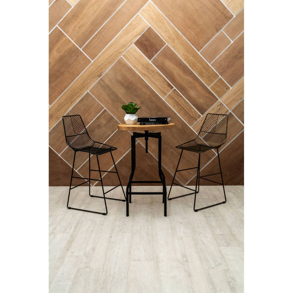 A bar table with a smooth ash veneer top and a sleek black metal base, accompanied by two wire-frame bar stools, set against a geometric wooden wall design. A small potted plant and a decorative item are placed on the table, with light-colored flooring visible beneath