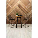 A bar table with a smooth ash veneer top and a sleek black metal base, accompanied by two wire-frame bar stools, set against a geometric wooden wall design. A small potted plant and a decorative item are placed on the table, with light-colored flooring visible beneath