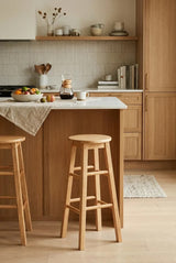 Tall bar stool made from natural tropical Hevea wood, featuring a simple and elegant design. The stool is positioned next to a kitchen island with a light-colored countertop, surrounded by a warm-toned kitchen setting that includes wooden cabinetry and decorative items on shelves. A textured table runner and some food items are visible on the countertop, enhancing the cozy atmosphere