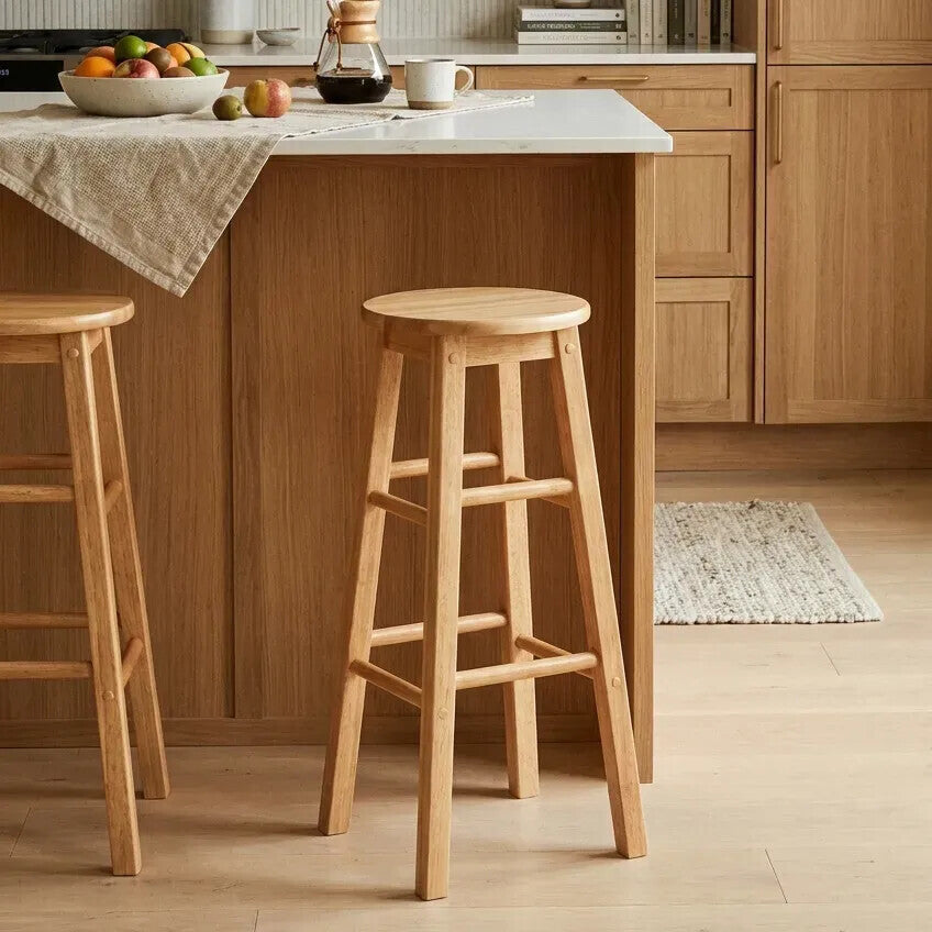 Tall bar stool made from natural tropical Hevea wood, featuring a round seat and four sturdy legs with a crossbar for support. The stool is positioned next to a kitchen island with a light-colored countertop, surrounded by a warm wooden cabinetry and a soft area rug on the floor. A bowl of fruit and other kitchen items are visible on the countertop
