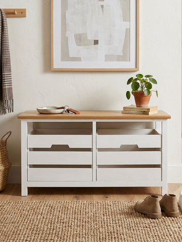 A white wooden bench with two spacious drawers and a natural wood top, placed against a light-colored wall. A potted plant sits on the bench, and there are decorative items and a framed artwork in the background. The floor is covered with a textured beige rug