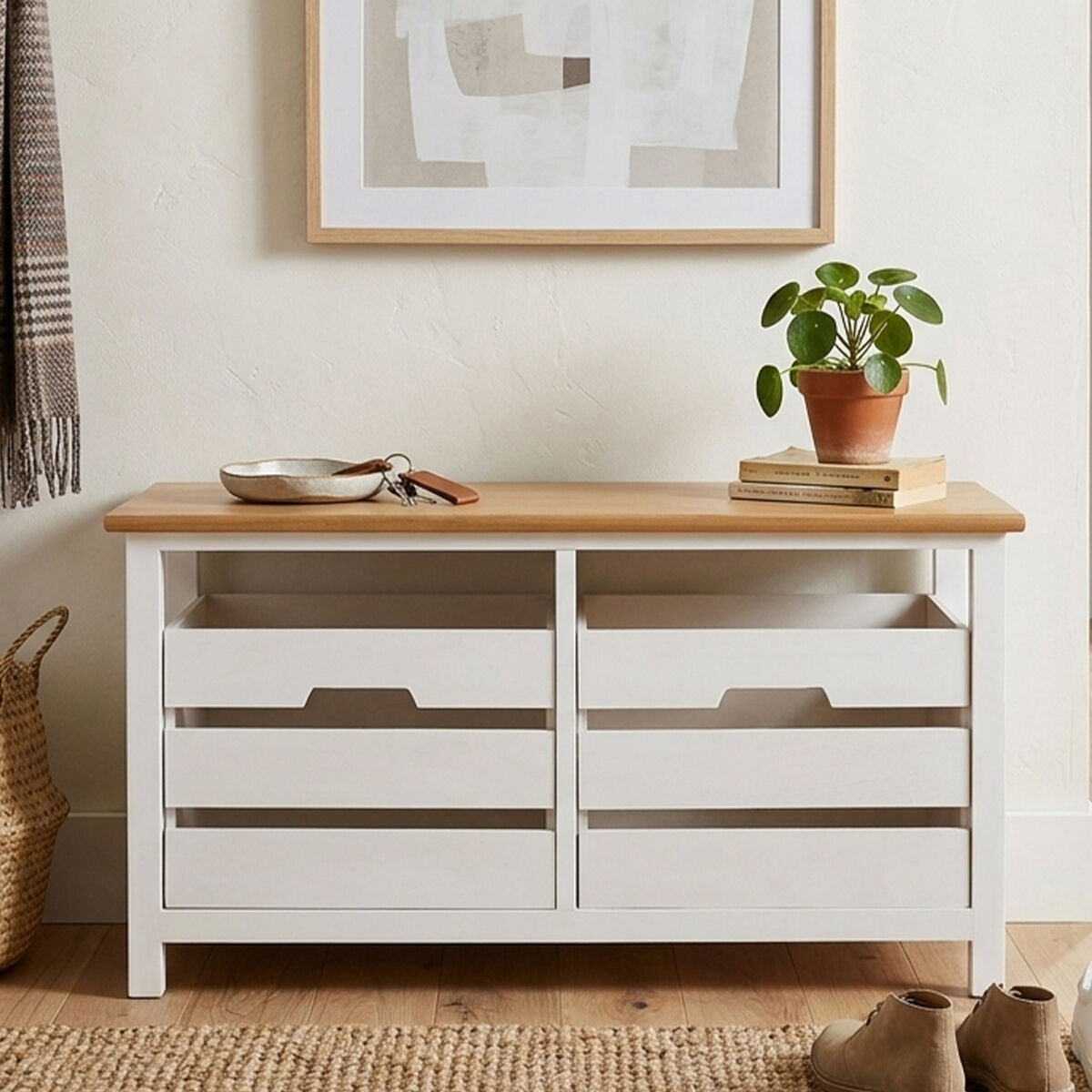 A white wooden bench with a natural wood top, featuring two spacious drawers below. The bench is placed against a light-colored wall, with a potted plant on top and a framed picture hanging above it. A woven basket is visible on the floor beside the bench