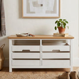 A white wooden bench with a natural wood top, featuring two spacious drawers below. The bench is placed against a light-colored wall, with a potted plant on top and a framed picture hanging above it. A woven basket is visible on the floor beside the bench
