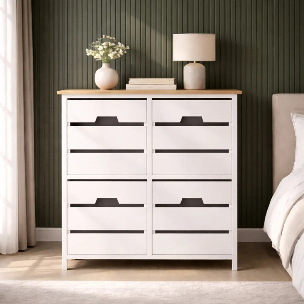A white four-drawer chest with a natural wood top, featuring sleek black handles, positioned against a green wall. A small vase with flowers and a lamp are placed on top of the chest, with a soft rug visible on the floor in front of it