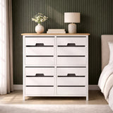 A white four-drawer chest with a natural wood top, featuring sleek black handles, positioned against a green wall. A small vase with flowers and a lamp are placed on top of the chest, with a soft rug visible on the floor in front of it