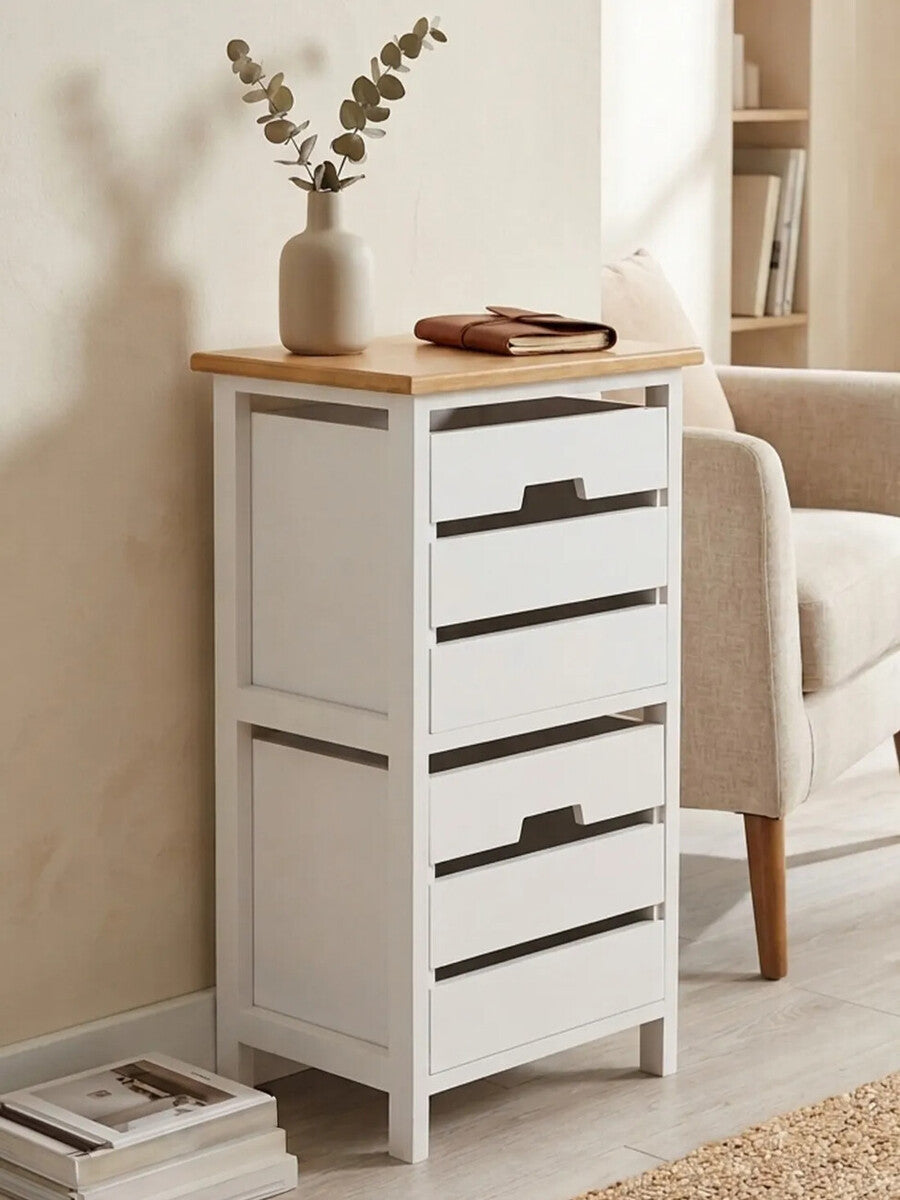A white two-drawer chest with a natural wood top, featuring sleek black handles, positioned against a light-colored wall. A small vase with dried flowers sits on top, and a cozy armchair is visible in the background