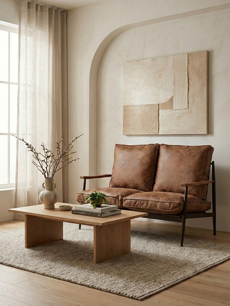 Brown leather effect 2 seater sofa with plush cushions, positioned in a cozy living room setting. A wooden coffee table with a small plant and decorative items sits in front of the sofa, while a textured area rug covers the floor. Natural light filters through sheer curtains, illuminating the space, which features a minimalist wall art piece in the background