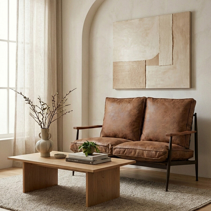 Brown leather effect 2 seater sofa with plush cushions, positioned in a cozy living room setting. A wooden coffee table with a small plant and decorative items sits in front of the sofa, while a textured area rug covers the floor. Natural light filters through sheer curtains, illuminating the space, which features a minimalist wall art piece in the background