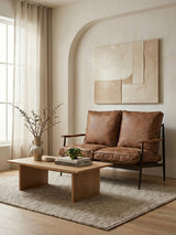 Brown leather effect 2 seater sofa with plush cushions, positioned in a cozy living room setting. A wooden coffee table with a small plant and decorative items sits in front of the sofa, while a textured area rug covers the floor. Natural light filters through sheer curtains, illuminating the space, which features a minimalist wall art piece in the background