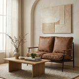 Brown leather effect two-seater sofa with plush cushions, set in a cozy living room. A wooden coffee table with decorative items sits in front of the sofa, and a large abstract wall art piece hangs above. Natural light filters through sheer curtains, illuminating the warm tones of the space