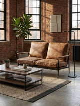 Brown leather effect two-seater sofa with plush cushions, set against a backdrop of exposed brick walls and large windows. A coffee table with a black metal frame and wooden top sits in front of the sofa, adorned with decorative items. A potted plant adds a touch of greenery to the industrial-style room, which features a textured area rug on the floor
