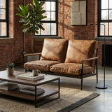 A brown leather effect two-seater sofa with plush cushions, set against a backdrop of exposed brick walls and large windows. A modern coffee table with stacked books and a decorative plant sits in front of the sofa, while a floor lamp stands nearby, illuminating the cozy space