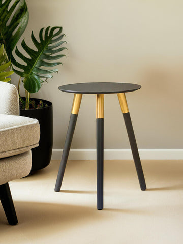 Round side table with a dark gray top and three tapered legs, two of which are black and one is natural wood. The table is positioned next to a light-colored sofa and a large green plant in a black pot is visible in the background. The setting features neutral wall colors and a soft floor