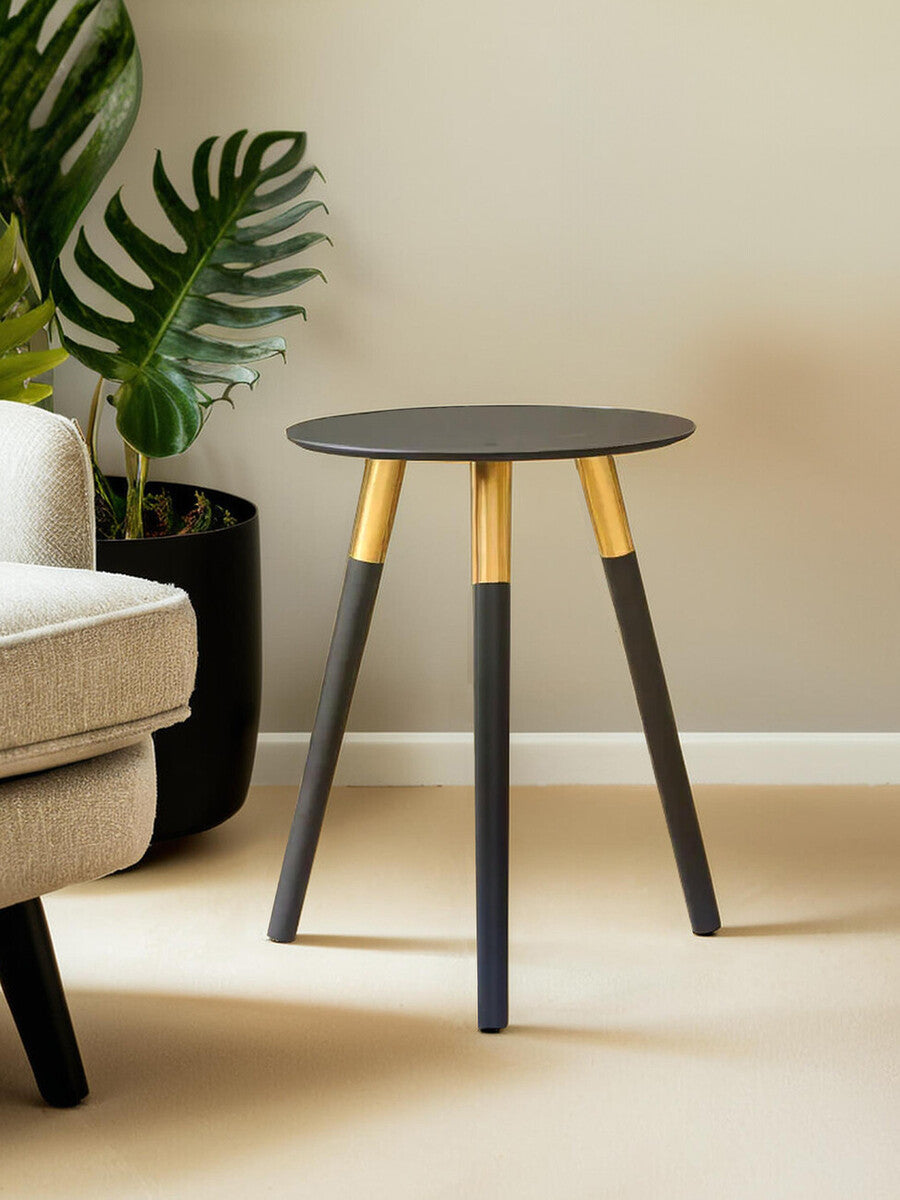 Round side table with a dark gray top and three tapered legs, two of which are black and one is natural wood. The table is positioned next to a light-colored sofa and a large green plant in a black pot is visible in the background. The setting features neutral wall colors and a soft floor