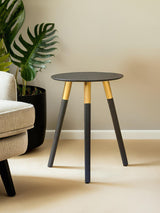 Round side table with a dark gray top and three tapered legs, two of which are black and one is natural wood. The table is positioned next to a light-colored sofa and a large green plant in a black pot is visible in the background. The setting features neutral wall colors and a soft floor
