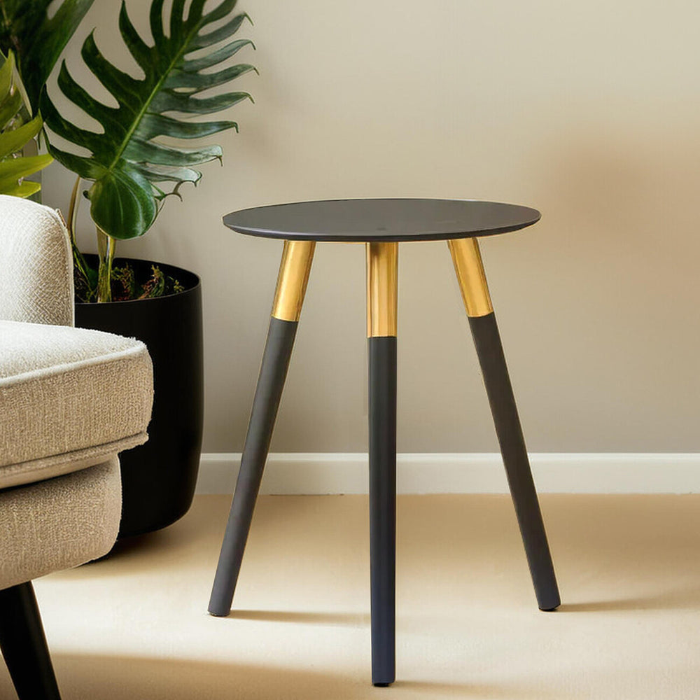 Round side table with a dark gray top and three legs, two of which are black and one is gold. The table is positioned next to a light-colored sofa and a potted plant in the background, set against a neutral wall