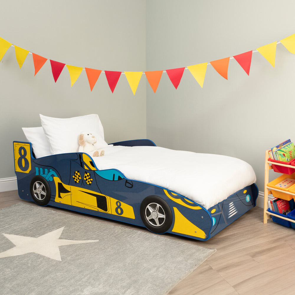 A colorful kids racing car bed designed in blue and yellow, featuring a sleek car shape with racing details. The bed is positioned against a light-colored wall, with a white mattress and pillows on top. A playful star-patterned rug is underneath, and a bright banner of triangular flags hangs above. A small storage unit filled with toys is visible in the corner