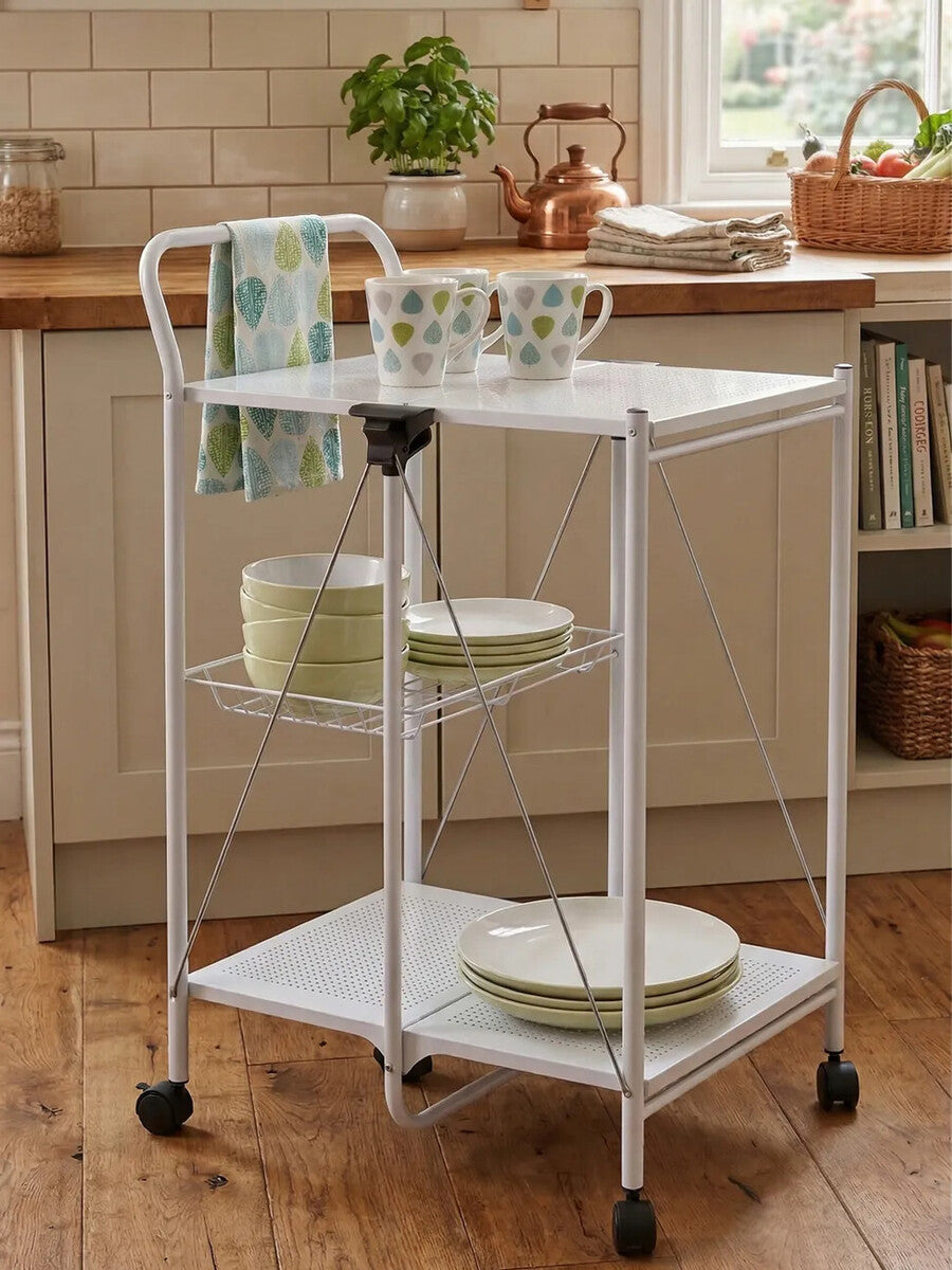 A white rectangular folding kitchen trolley with two shelves, featuring a top surface holding two mugs and a stack of plates. The trolley has a handle on one side and is mounted on wheels for easy mobility. The background shows a cozy kitchen setting with wooden cabinets and a warm, inviting atmosphere