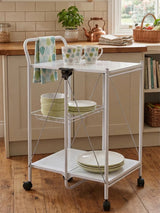 A white rectangular folding kitchen trolley with two shelves, featuring a top surface holding two mugs and a stack of plates. The trolley has a handle on one side and is mounted on wheels for easy mobility. The background shows a cozy kitchen setting with wooden cabinets and a warm, inviting atmosphere
