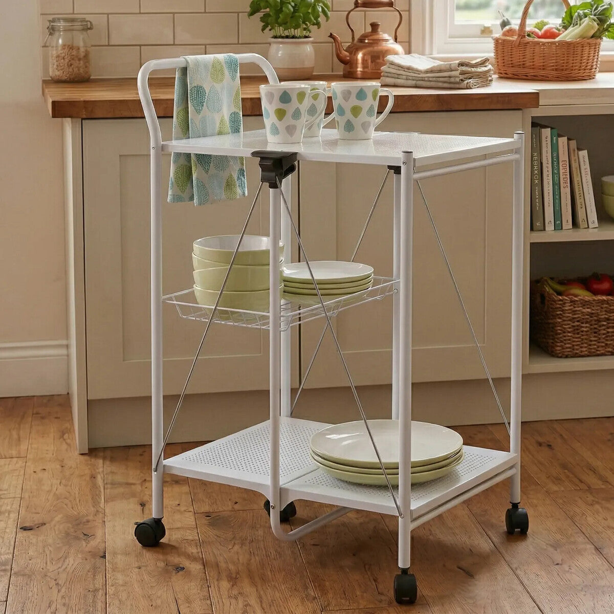 White rectangular folding kitchen trolley with three shelves, featuring a top surface holding bowls and cups, a towel hanging on the side, and four wheels for mobility, set against a wooden kitchen backdrop with natural light